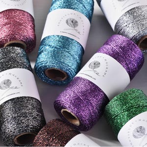 Può includere: Diverse bobine di cordino in macramè metallizzato in vari colori, tra cui rosa, blu, argento, viola, verde e marrone. Ogni bobina presenta un'etichetta di carta bianca con il testo Knotitude Macrame e A Happy Handmade Life.