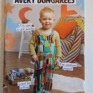 May include: A sewing pattern envelope titled AVERY DUNGAREES featuring a child wearing colourful, multi-patterned dungarees over a mustard yellow long-sleeve shirt. The cover includes text stating Fits ages 6-24 months, Fun weekend project!, and Great for gifting.