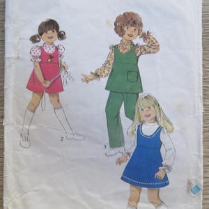Pode incluir: Um envelope de molde de costura vintage Style, número 4265, para roupas de criança tamanho 4. A frente apresenta ilustrações de três crianças vestindo um vestido rosa, um macacão verde e um vestido azul, combinados com camisas de manga comprida e meias.