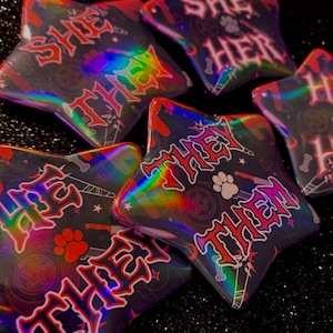 May include: Five holographic star-shaped buttons with different pronouns written on them. The pronouns are "She/Her", "They/Them", "She/They", "He/Him", and "They/Them". The buttons are on a black sparkly background.
