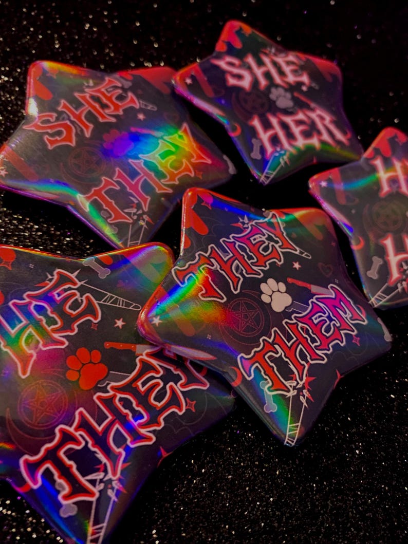 May include: Five holographic star-shaped buttons with different pronouns written on them. The pronouns are "She/Her", "They/Them", "She/They", "He/Him", and "They/Them". The buttons are on a black sparkly background.