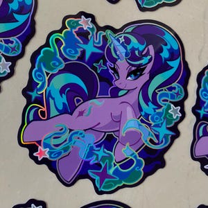 May include: A holographic sticker featuring a purple unicorn with a blue and green mane and tail. The unicorn is surrounded by stars and clouds. The sticker has a glossy finish.