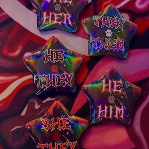 May include: Five star-shaped buttons with a holographic finish. Each button features a different pronoun set in a gothic font: "She Her", "They Them", "He They", "He Him", and "She They".