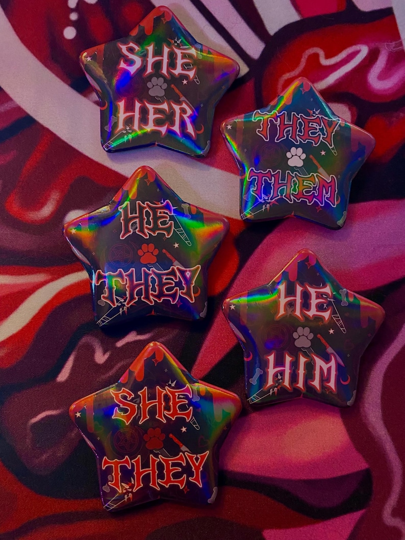May include: Five star-shaped buttons with a holographic finish. Each button features a different pronoun set in a gothic font: "She Her", "They Them", "He They", "He Him", and "She They".