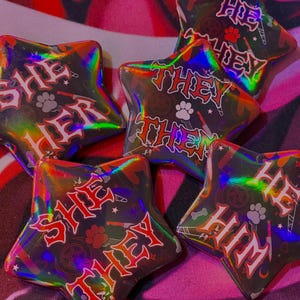 May include: Five iridescent star-shaped pins with black backgrounds and red text. The pins read "She Her", "They Them", "He Him", "She They", and "Hoe They".