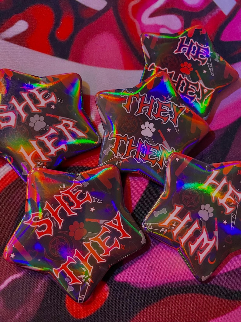 May include: Five iridescent star-shaped pins with black backgrounds and red text. The pins read "She Her", "They Them", "He Him", "She They", and "Hoe They".