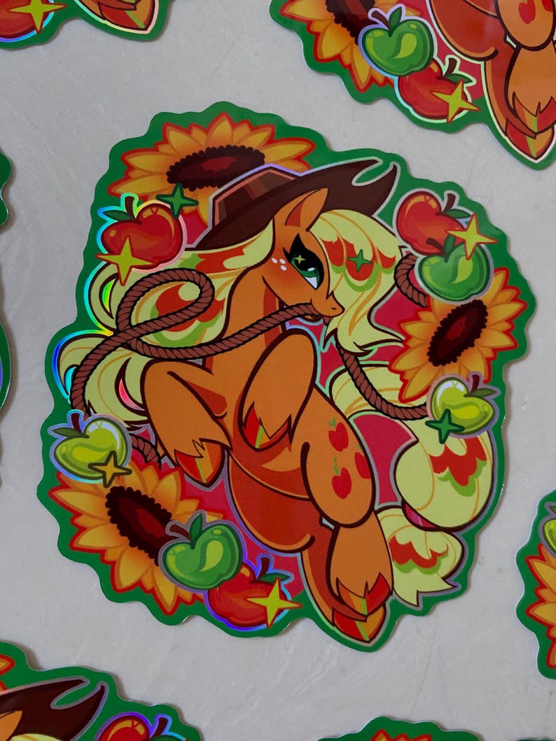 May include: A holographic sticker featuring a cartoon pony with a cowboy hat, a lasso, and apples. The pony is orange and yellow with a pink mane and tail. The background is green with sunflowers and stars.