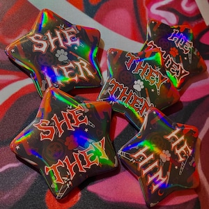 May include: Five star-shaped buttons with a black background and holographic rainbow effect. The buttons feature different phrases in red and white lettering: "She Her", "They Them", "She They", "He Him", and "He They".