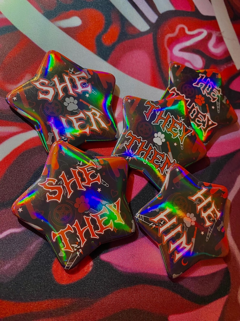 May include: Five star-shaped buttons with a black background and holographic rainbow effect. The buttons feature different phrases in red and white lettering: "She Her", "They Them", "She They", "He Him", and "He They".