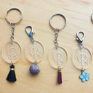 Peut inclure: Cinq porte-clés avec des breloques rondes transparentes ornées d'un logo blanc. Chaque porte-clés a un pompon de couleur différente : rouge, noir, gris, marron et turquoise.