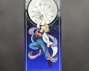 May include: A bookmark featuring a cartoon illustration of a person in a white lab coat and blue jeans, reaching up to touch a crescent moon with stars. The person is surrounded by a rainbow-coloured ribbon and is floating above a blue and white planet.
