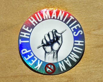 May include: Round sticker with a holographic rainbow border. The sticker features a black and white illustration of a clenched fist holding a pencil, with the words "KEEP THE HUMANITIES HUMAN" in a circular design. A red circle with a line through it is at the bottom.