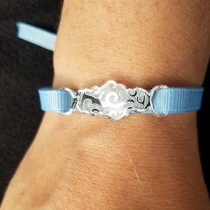 May include: A light blue ribbon bracelet with a silver charm featuring a stylized cloud design.