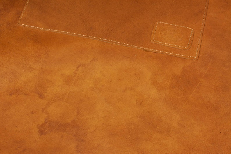 May include: Close-up of a brown leather surface with a stitched seam and a small rectangular patch of leather. The leather has a worn, distressed look.