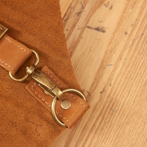 May include: Close-up of a brown leather strap with a brass buckle and a metal hook. The strap is attached to a larger piece of leather.