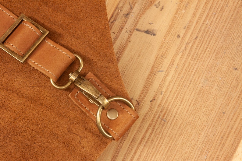 May include: Close-up of a brown leather strap with a brass buckle and a metal hook. The strap is attached to a larger piece of leather.
