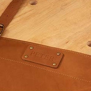 May include: A brown leather apron with adjustable straps and a metal buckle. The apron has a rectangular leather patch with the word "PETISR" embossed on it.