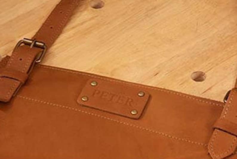 May include: A brown leather apron with adjustable straps and a metal buckle. The apron has a rectangular leather patch with the word "PETISR" embossed on it.