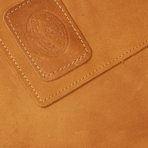 May include: Close-up of a brown leather surface with a stitched seam. A smaller rectangular piece of leather with a stitched border is on top of the larger piece. The smaller piece has a circular embossed design.
