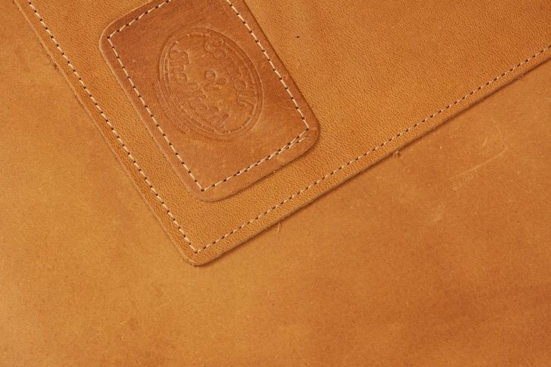 May include: Close-up of a brown leather surface with a stitched seam. A smaller rectangular piece of leather with a stitched border is on top of the larger piece. The smaller piece has a circular embossed design.