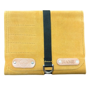 May include: A tan leather tool roll with a black strap and a silver D-ring. The roll has multiple slots for tools and a leather patch with the word "NAME" embossed on it.