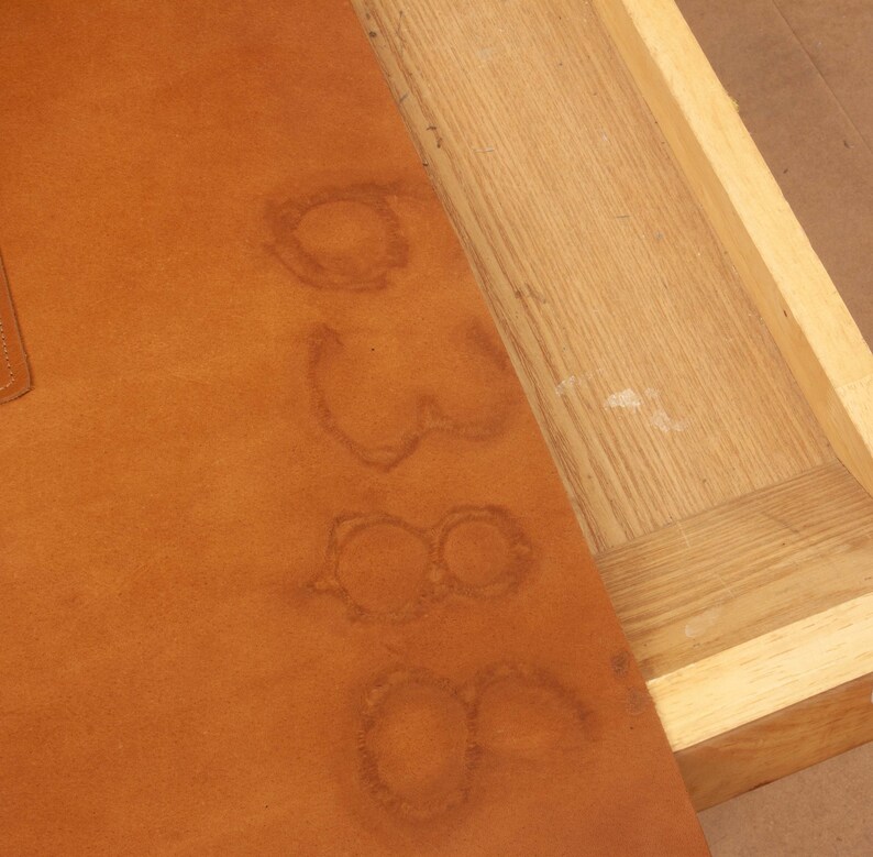 May include: A close-up of a brown leather surface with several watermarks in the shape of circles. The leather is part of a drawer or box, with the wooden edge of the drawer visible.