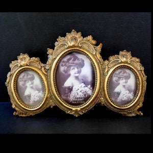 Puede incluir: Un marco de fotos triple en oro antiguo con un diseño ornamentado. Cuenta con tres aberturas ovaladas, cada una con un retrato en sepia. El marco central está etiquetado como "Photo frame 9cm x 13cm".