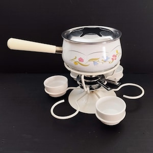 Op de afbeelding: Witte keramische fonduepot met een bloemenmotief en een witte metalen standaard met vier dipschalen.