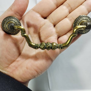 Op de afbeelding: Een messing ladegreep met sierlijke details en een decoratief ontwerp. De greep heeft twee ronde uiteinden met een gebogen verbindend stuk.