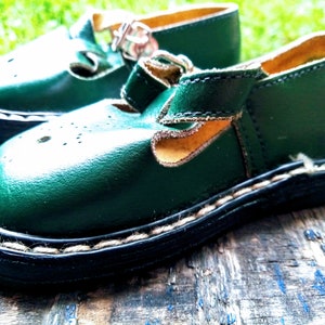 Pode incluir: Um par de sapatos de couro verde para crian&ccedil;as com fecho de fivela e borda costurada. Os sapatos t&ecirc;m um forro de couro castanho e um detalhe de costura branca em volta da sola.