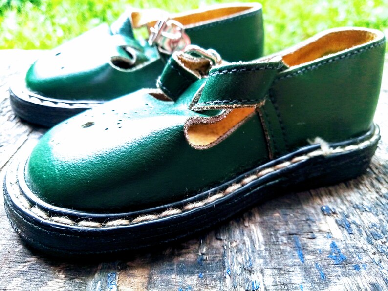 Pode incluir: Um par de sapatos de couro verde para crian&ccedil;as com fecho de fivela e borda costurada. Os sapatos t&ecirc;m um forro de couro castanho e um detalhe de costura branca em volta da sola.