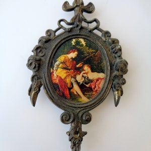 May include: An antique oval-shaped wall hanging with a detailed bronze frame. The frame surrounds a small, colorful print depicting two figures in a pastoral scene. The frame has ornate detailing and a dark, aged finish.