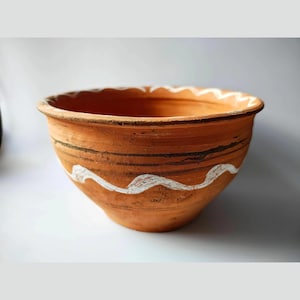 Könnte beinhalten: Eine Terrakotta-Schüssel mit breitem Rand und rundem Körper. Die Schüssel hat ein wellenförmiges, weißes Design um die Mitte und den Rand. Die Schüssel ist unglasiert und hat ein rustikales, handgemachtes Aussehen.