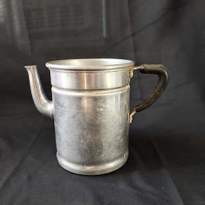 Puede incluir: Una cafetera de aluminio plateado con un asa negra. La cafetera tiene un cuerpo redondeado y un pico en el lateral.