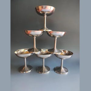 Könnte beinhalten: Eine Pyramide aus sechs silbernen Champagner-Coupe-Gläsern. Die Gläser haben ein klassisches Design mit einer breiten, flachen Schale und einem langen Stiel. Die Gläser sind in einer gestapelten Formation angeordnet und erzeugen eine ansprechende Darstellung.