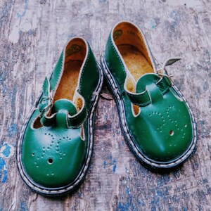 Pode incluir: Um par de sapatos de couro verde para crian&ccedil;as com fecho de fivela. Os sapatos t&ecirc;m uma borda branca em volta da sola e um pequeno padr&atilde;o de furos no couro.