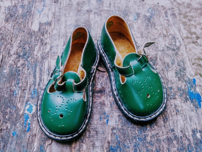 Pode incluir: Um par de sapatos de couro verde para crian&ccedil;as com fecho de fivela. Os sapatos t&ecirc;m uma borda branca em volta da sola e um pequeno padr&atilde;o de furos no couro.