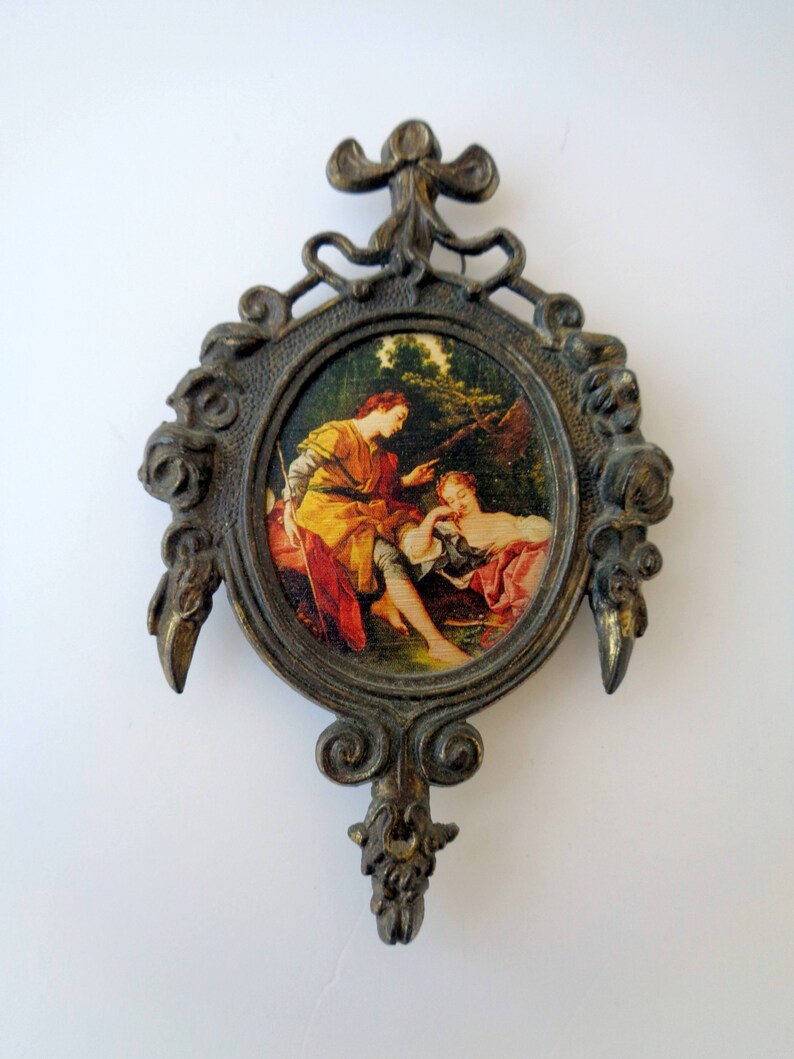 May include: Ornate, antique-style wall hanging with a circular painting depicting two figures in a garden setting. The frame is bronze-colored with decorative scrollwork and a bow-shaped top. The painting features warm colors.