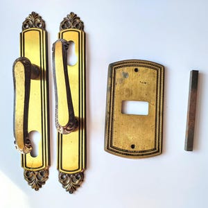 May include: An antique brass door hardware set. It includes two ornate door handle plates with handles, a keyhole cover, and a small rectangular piece. The hardware has a gold finish with dark brown accents and decorative detailing.
