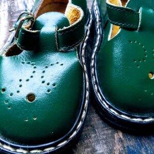 Pode incluir: Um par de sapatos de couro verde para crian&ccedil;as com fecho de fivela e costuras brancas ao redor da borda.