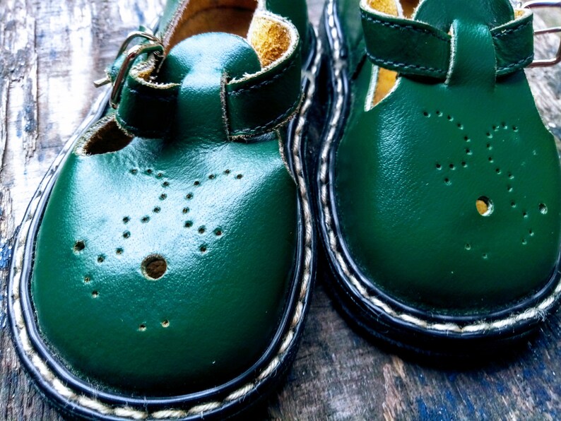 Pode incluir: Um par de sapatos de couro verde para crian&ccedil;as com fecho de fivela e costuras brancas ao redor da borda.