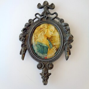 May include: An antique-style, oval-shaped framed artwork. The frame is dark metal with ornate detailing. The artwork depicts a woman in a blue dress with her hands clasped in prayer. The background is a blend of yellow and brown tones.