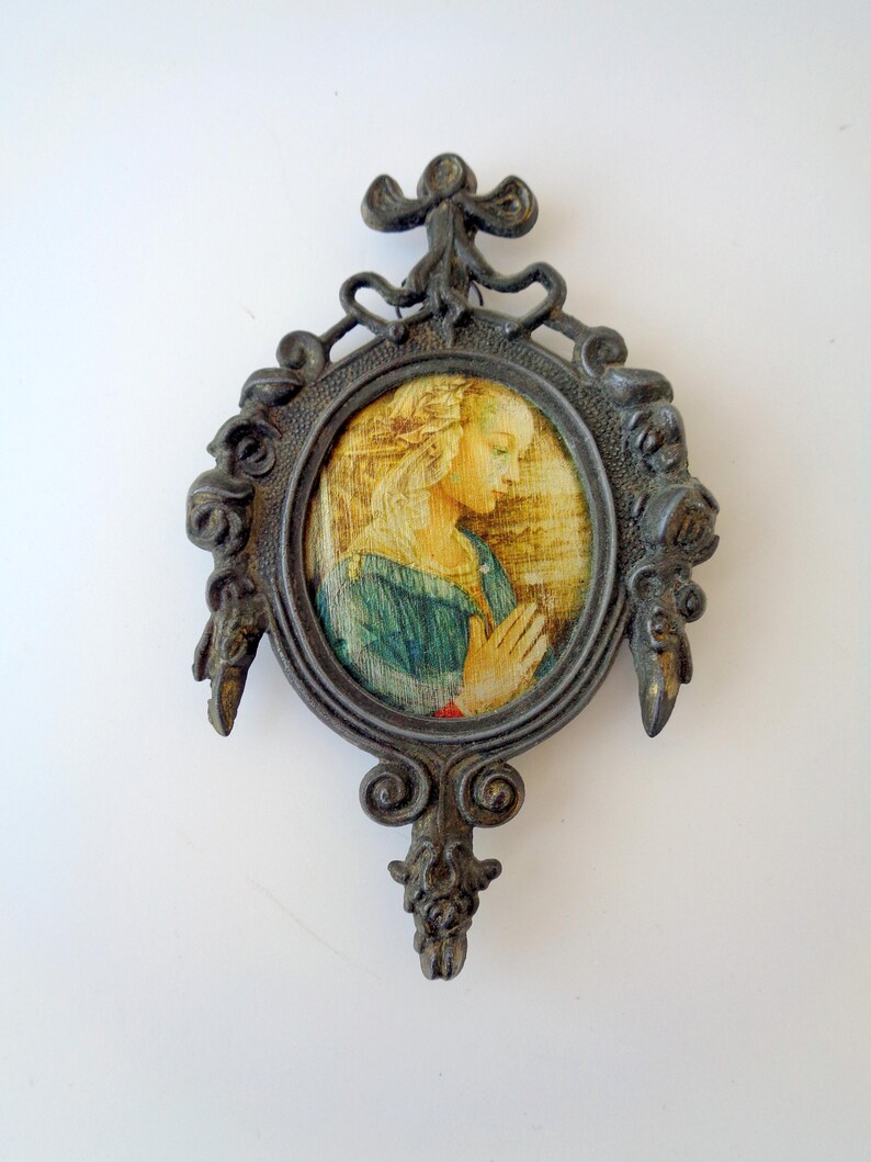 May include: An antique-style, oval-shaped framed artwork. The frame is dark metal with ornate detailing. The artwork depicts a woman in a blue dress with her hands clasped in prayer. The background is a blend of yellow and brown tones.