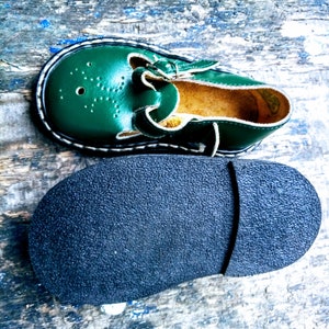 Pode incluir: Um par de sapatos de couro verde para crian&ccedil;as com fecho de fivela. Os sapatos t&ecirc;m uma sola de borracha preta e um detalhe de costura branca ao redor da borda.