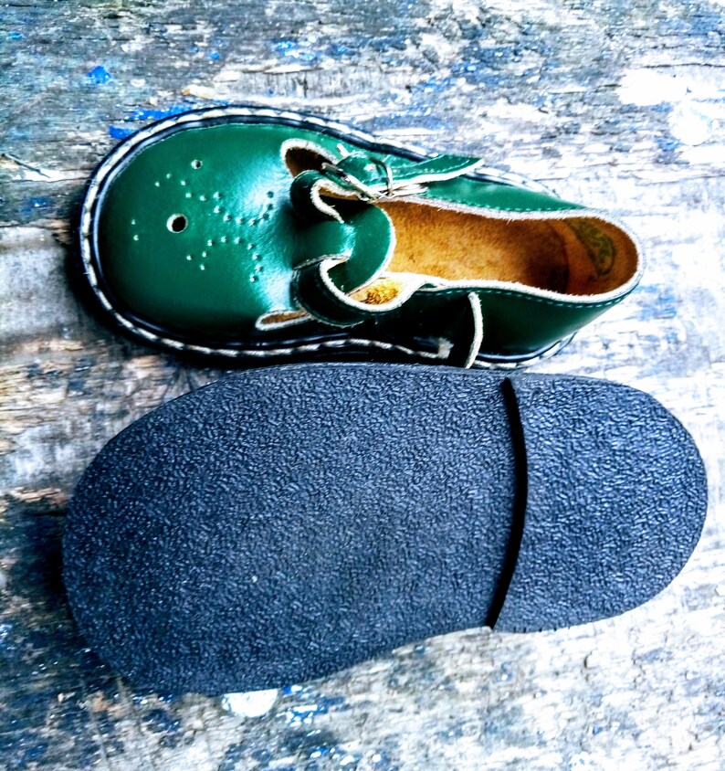 Pode incluir: Um par de sapatos de couro verde para crian&ccedil;as com fecho de fivela. Os sapatos t&ecirc;m uma sola de borracha preta e um detalhe de costura branca ao redor da borda.