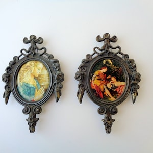 May include: Two antique oval picture frames with ornate bronze-colored metalwork. Each frame holds a small, colorful print of classical art. One depicts a woman in prayer, the other a couple.