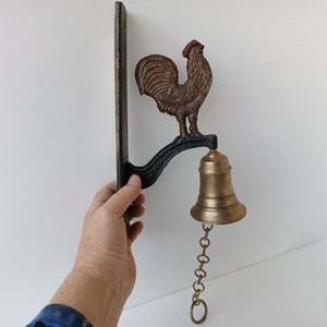 Puede incluir: Una campana de latón con una cadena cuelga de un soporte de metal negro con una figura de gallo. El gallo es marrón y rojo con un pico y ojos negros.