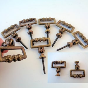 Könnte beinhalten: Acht antike Messing-Schubladengriffe mit verzierten Details. Jeder Griff hat eine rechteckige Form mit einem dekorativen Knopf in der Mitte. Die Griffe haben einen Vintage-Look und wären perfekt, um jedem Möbelstück einen Hauch von Eleganz zu verleihen.