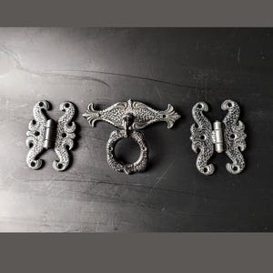 Può includere: Tre cerniere in metallo in stile antico con disegni decorativi. Le cerniere sono nere e hanno una finitura testurizzata. La cerniera centrale ha una maniglia ad anello.