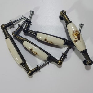 Peut inclure: Quatre boutons de cabinet de style antique avec un motif floral. Les boutons sont en céramique avec un fond blanc et des accents floraux bruns. Les boutons sont fixés à une base en laiton.